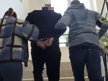 Funkcjonariusz z opaską na ramieniu i kobieta prowadzą w kajdankach mężczyznę po schodach wewnątrz budynku.